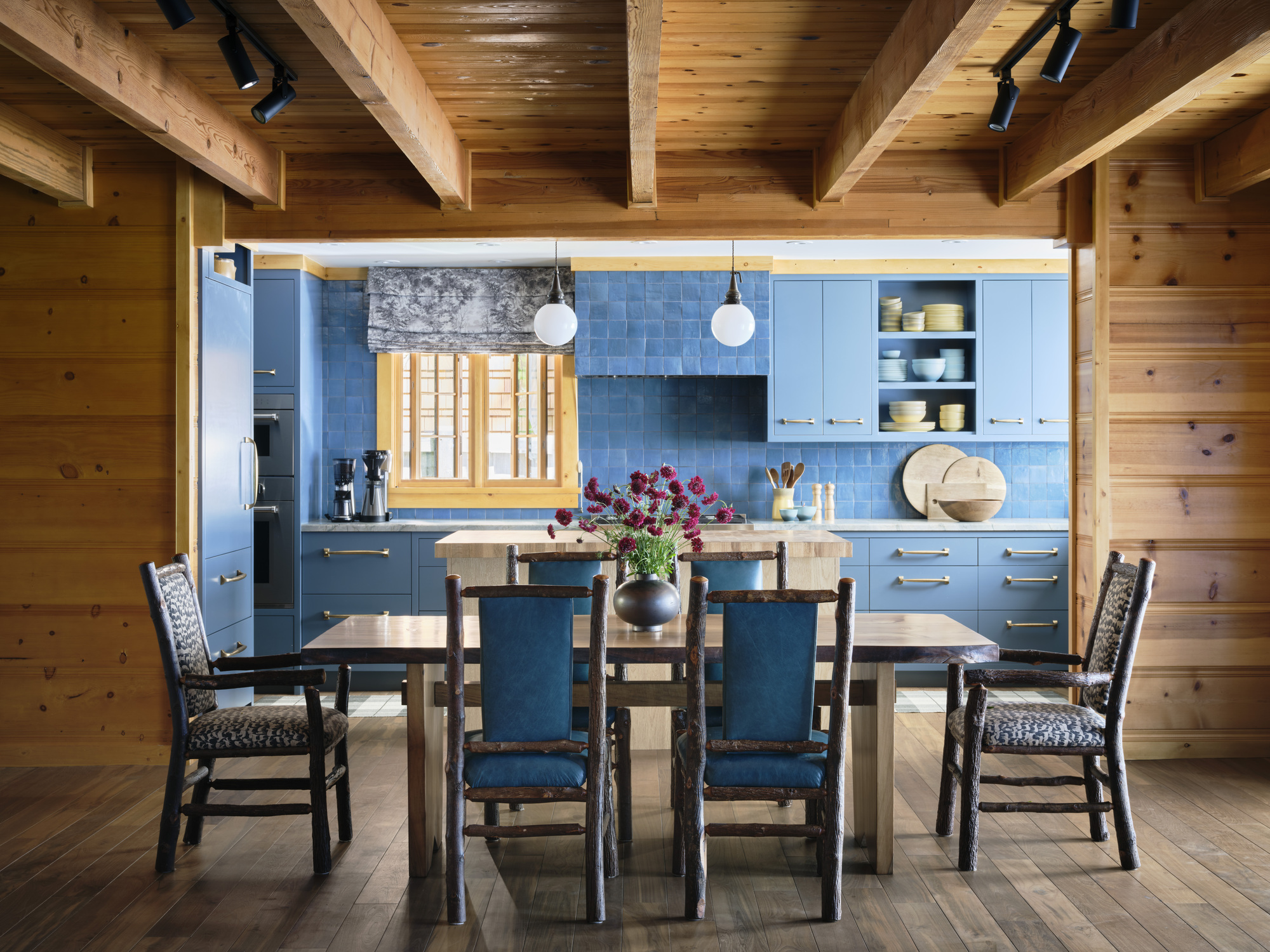 A dining room with wooden panelled walls and ceiling with a view into a kitchen with blue zellige tiles and blue cabinetry