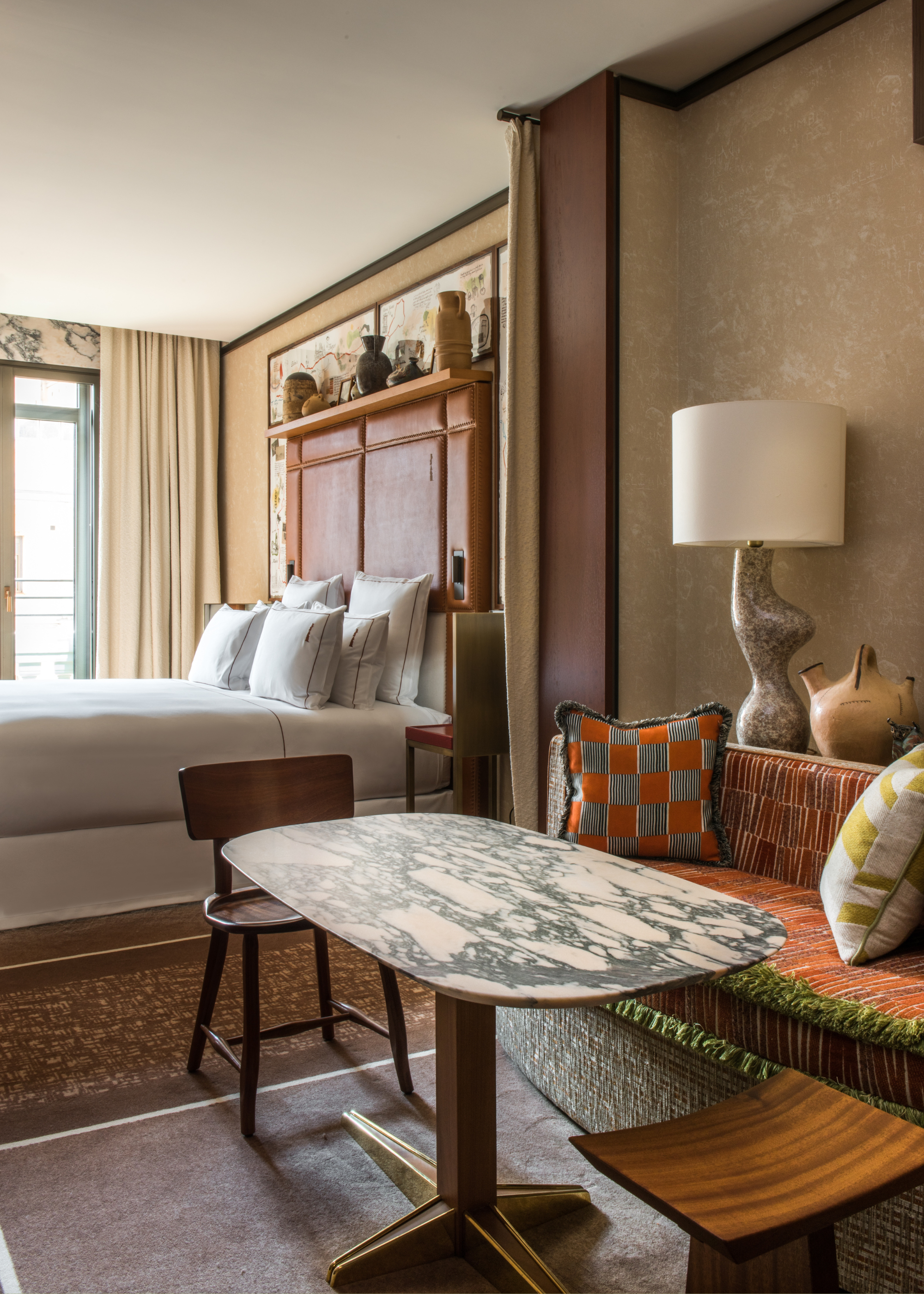 A bedroom with bright windows, a freshly made bed, a side sofa with a stone and wood table and mismatched chairs