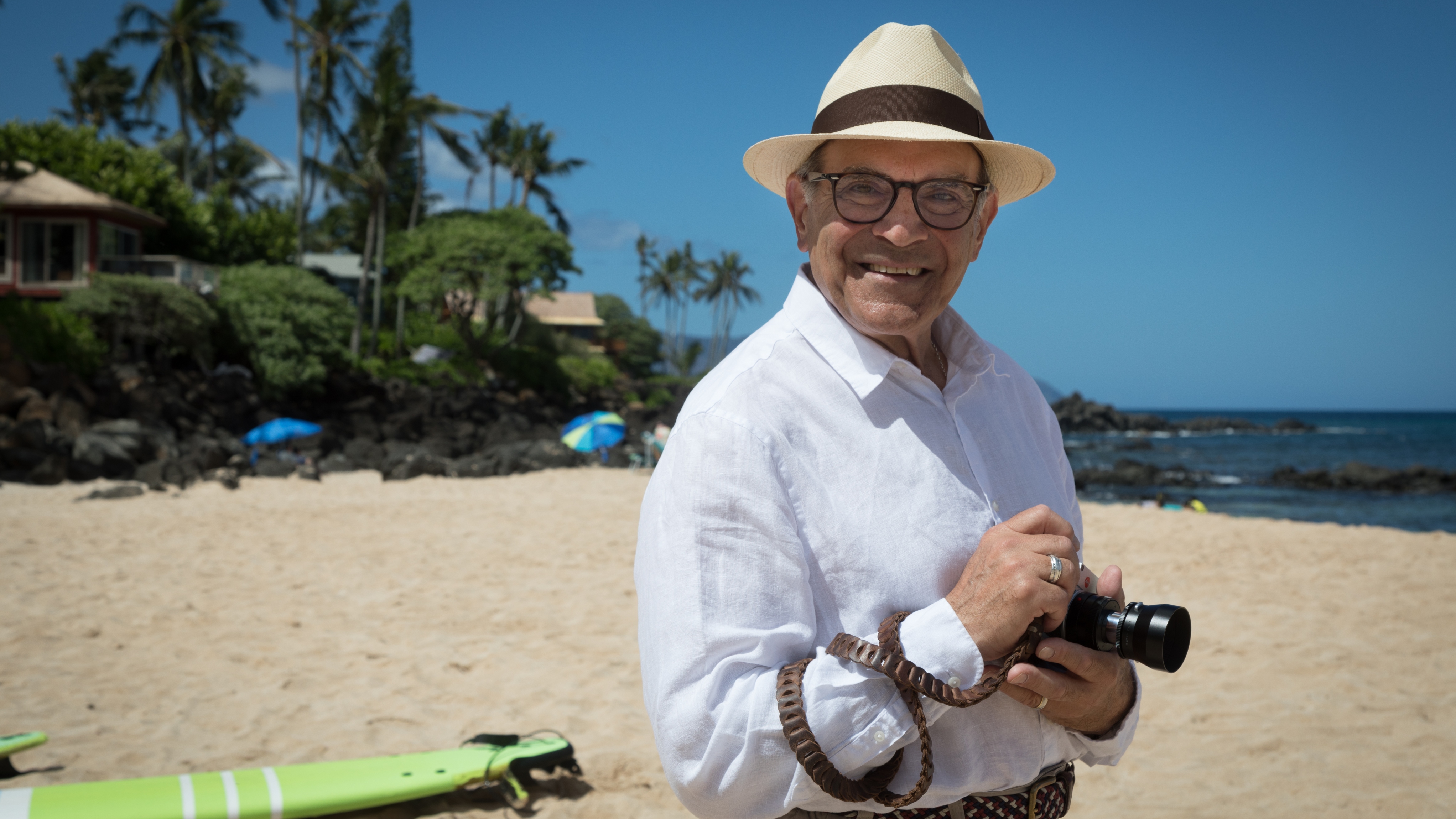 Sir David Suchet on a beach in Hawaii holding a camera and with a surfboard behind him.