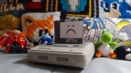 SNES (Super Famicom) console sitting on grey sofa with NES cart inserted and sad face attached to front.