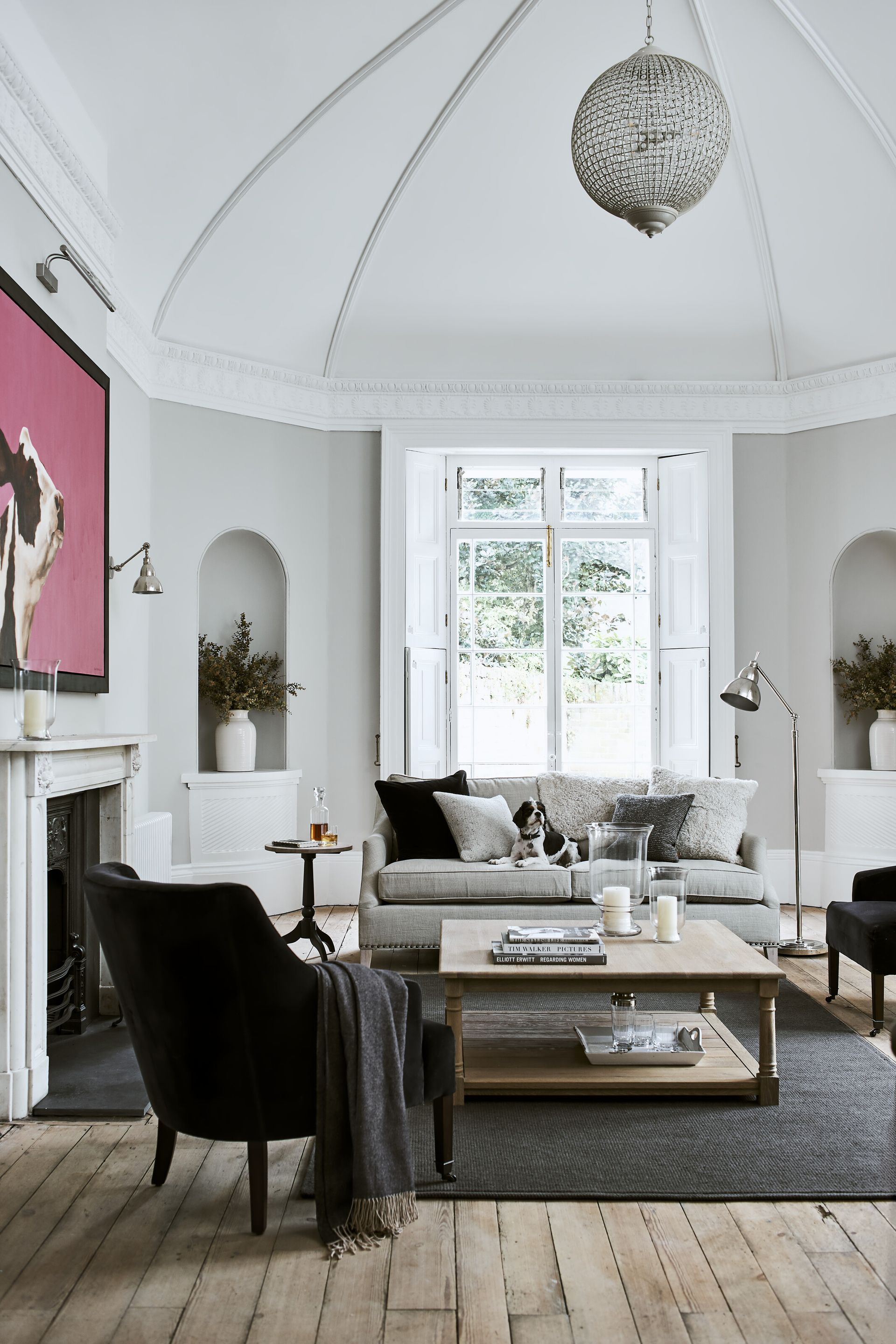 white and grey living room with vaulted ceiling, alcoves, grey sofa, wooden floorboards, artwork, chandelier, bay window