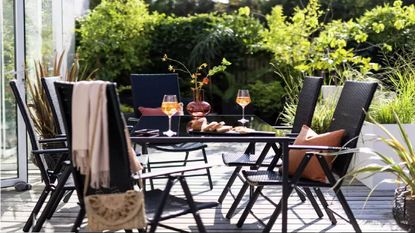 Black coloured rattan garden dining set with 6 chairs and glass table. Dining set is in sunny garden and the table has wine glasses and bread on it