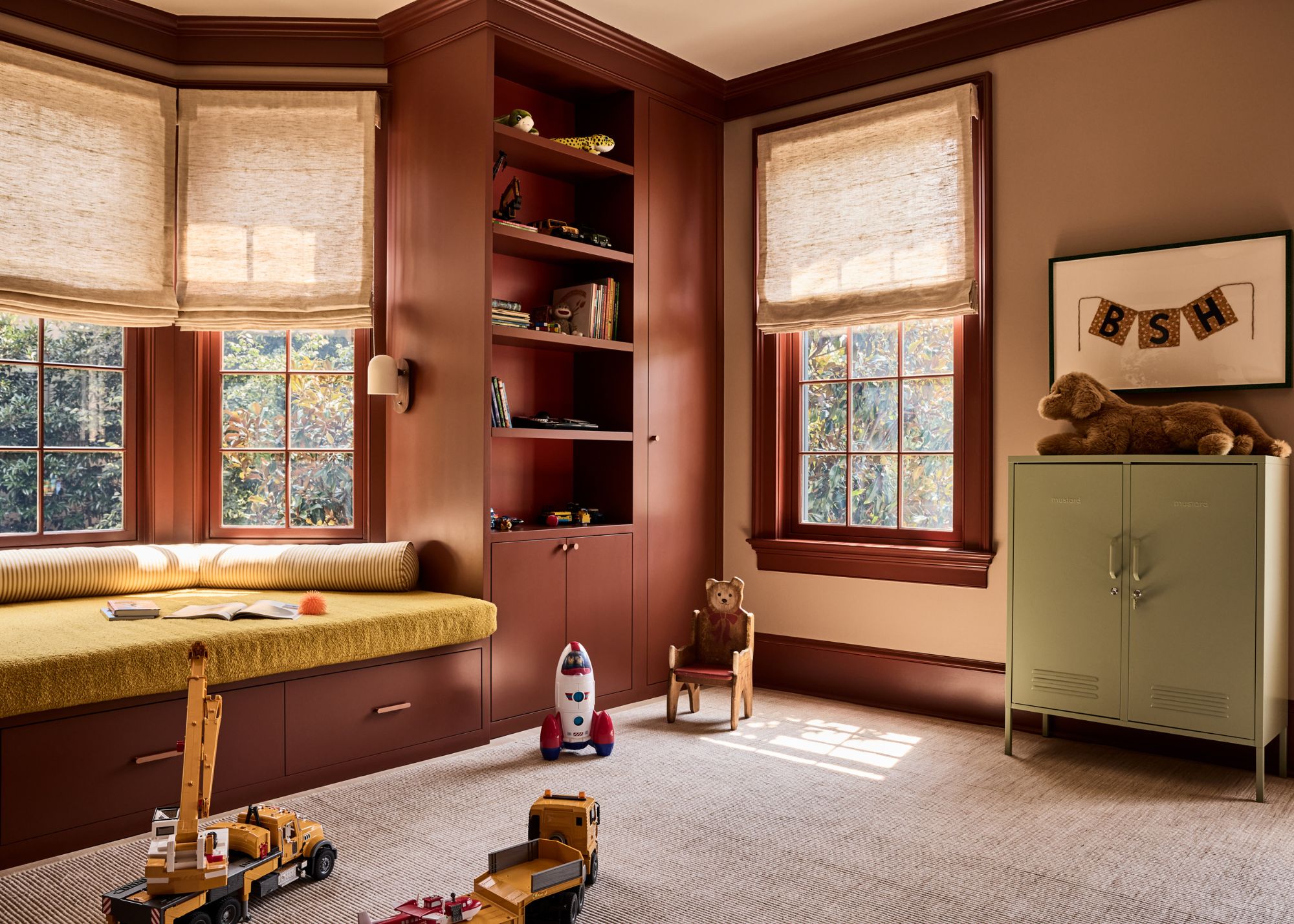 A kids' room with beige walls with brown-red trim and window casing, and full-height built-in shelving on the other wall. A bay window with a mustard bench seat and soft beige roman blinds.