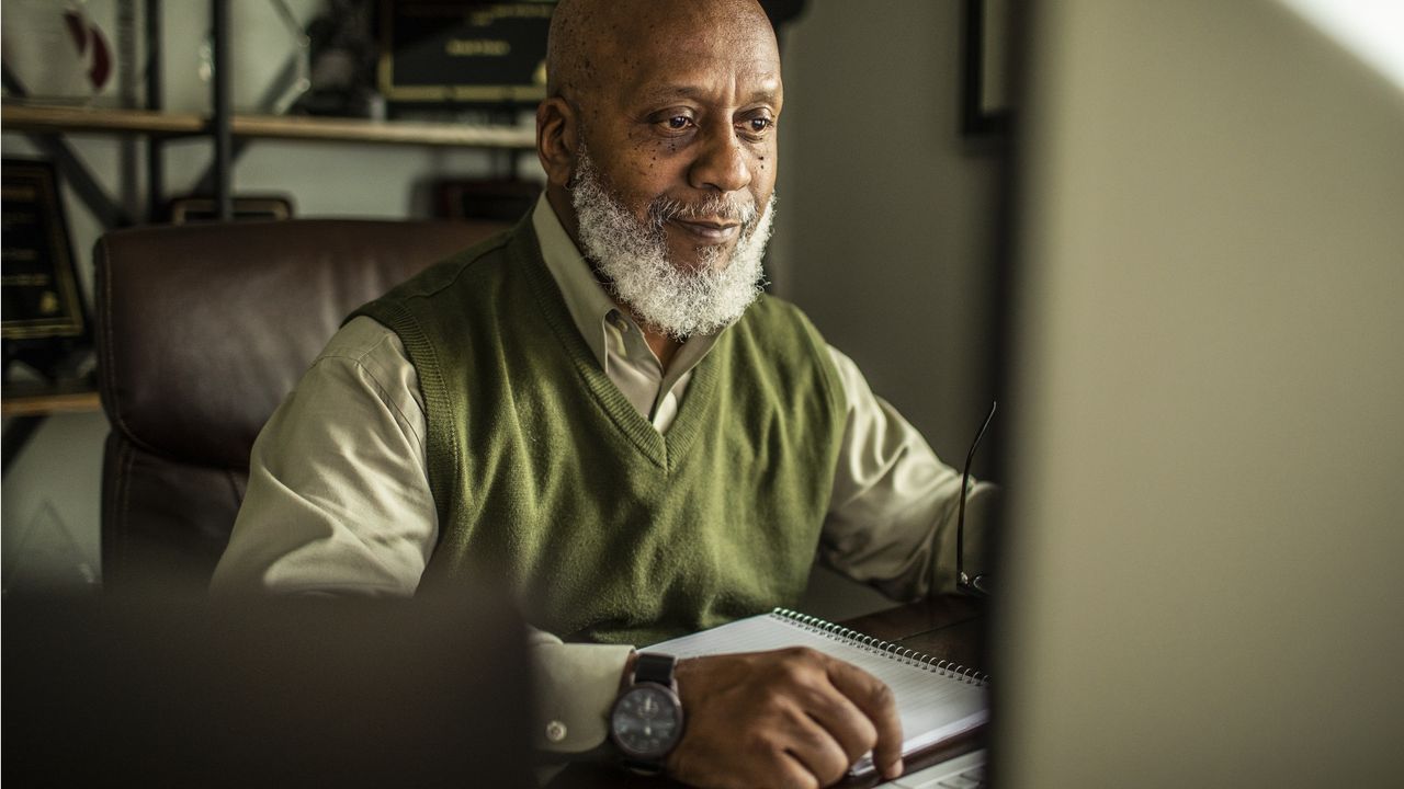 An older man sits at his computer in his home office.