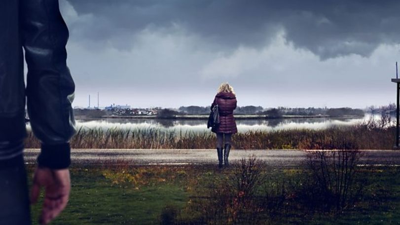 Sambre – Anatomy of a Crime still - woman standing by a river with man watching her from behind 