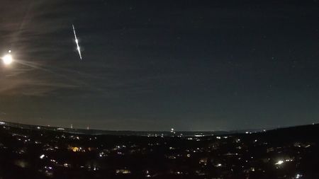A bright fireball meteor is shown streaking earthward through the night sky over the lights of a town. The bright reflected light of the moon is visible to its left.