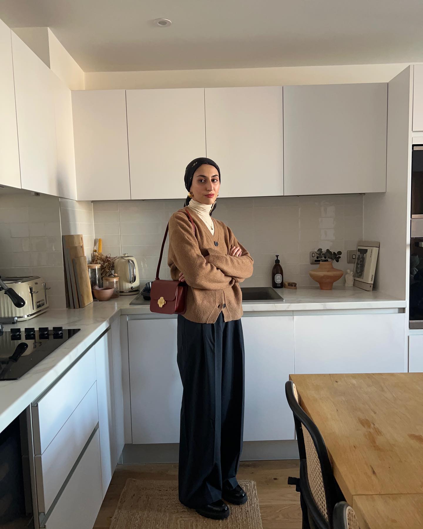 Woman standing in a kitchen wearing white turtleneck, brown cardigan, wide-leg trouser pants, burgundy shoulder bag