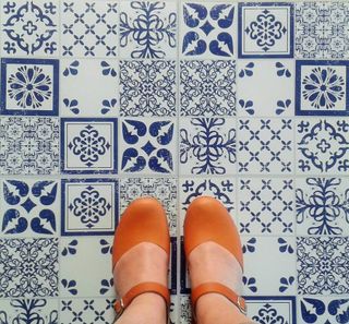 Azulejos Vinyl Floor Tiles in blue and white patterns, showing a birds eye view of a ladies feet standing on them wearing orange closed toe sandles