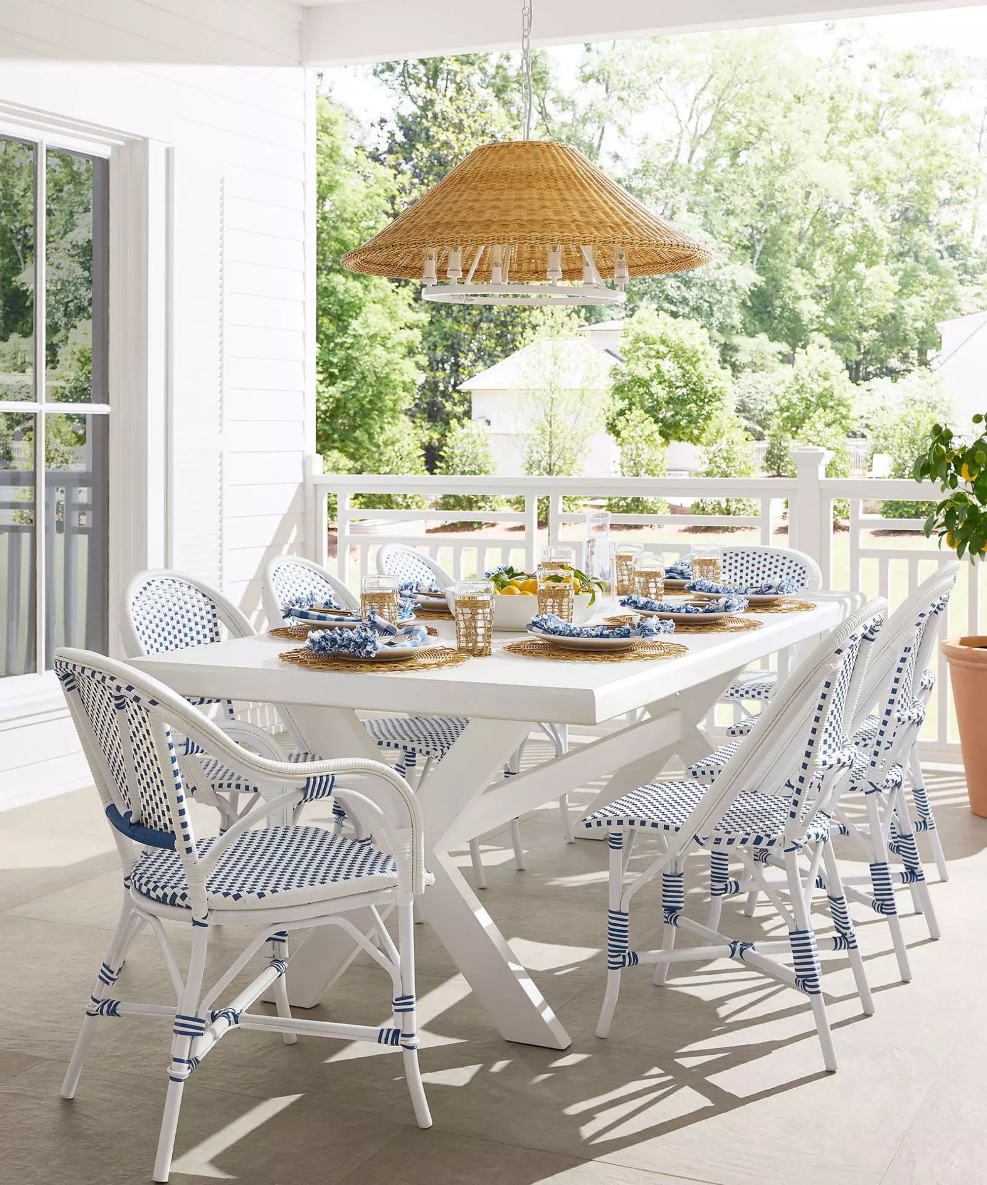 A sunlit covered porch featuring a white farmhouse-style dining table paired with blue and white woven bistro chairs. A large, conical rattan chandelier hangs above the set table, which is decorated with blue patterned linens and fresh lemons, overlooking a lush green backyard