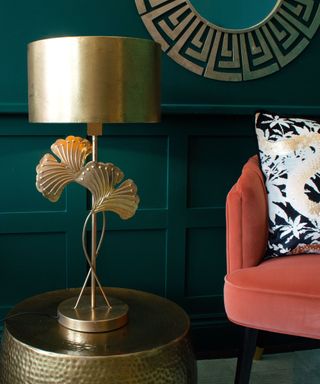 A gold lamp with a gold mirror and a coral chair next to it