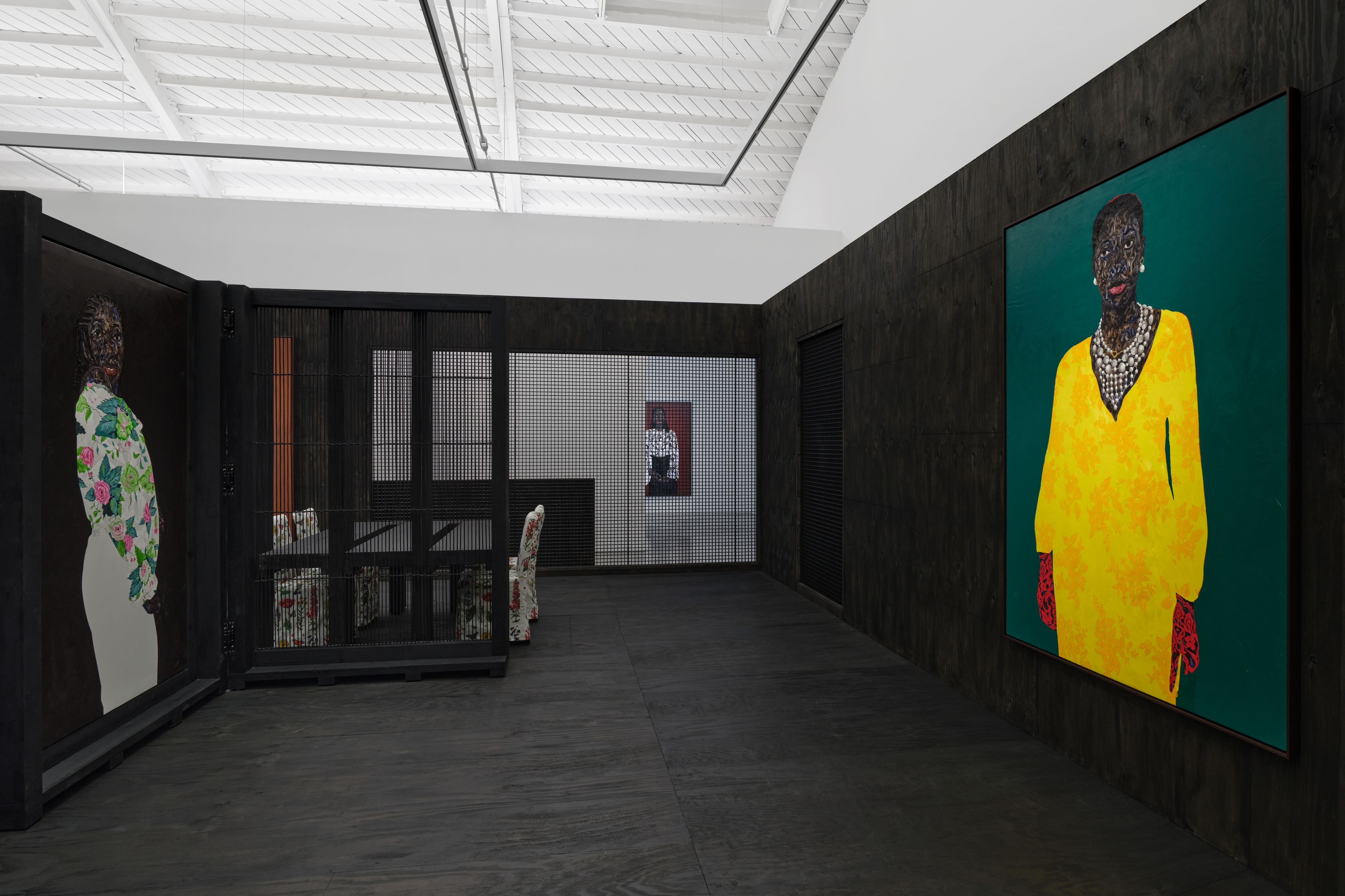 View of an art gallery installation with dark wood walls and multiple expressive portrait paintings. In the foreground, a large black metal structure with vertical bars frames additional paintings inside, while more portraits hang on the walls in the background.