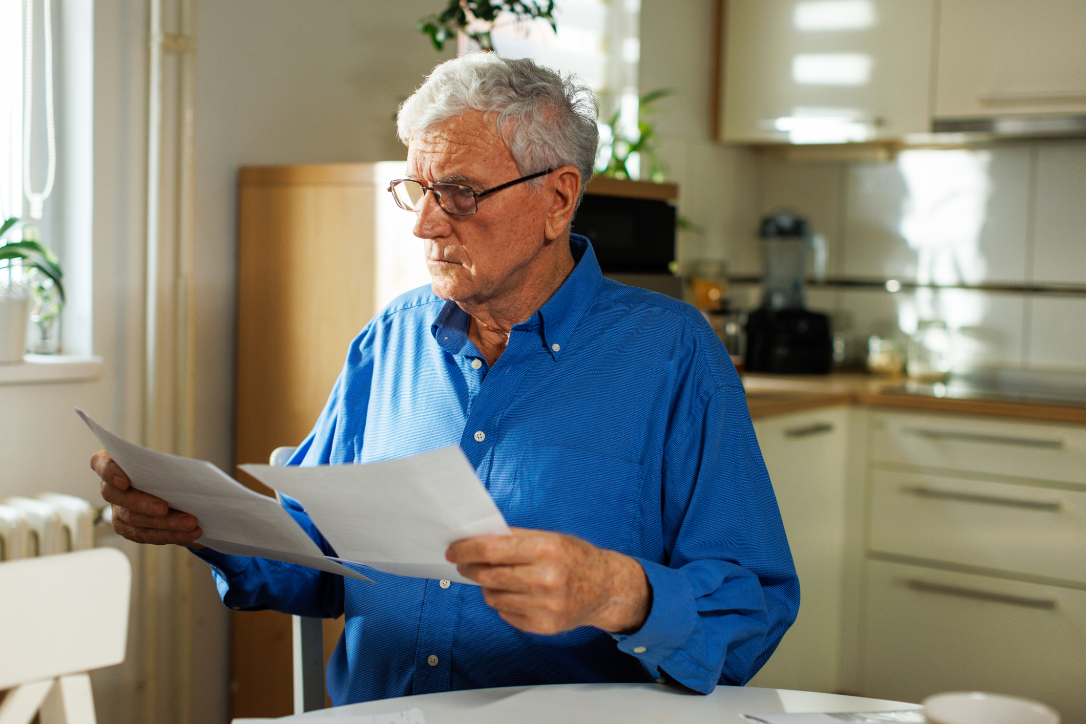 Pensioner looks at financial documents as he sits at kitchen table.