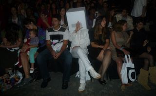 In the front row of the audience, a person in white wears a white box covering their head
