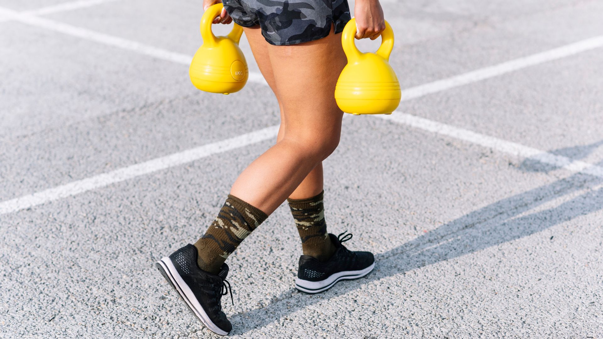 Woman doing farmer's carry exercise with two kettlebells, walking along tarmac pavement