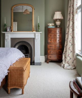 Classicly styled bedroom with cream carpet, fireplace, green painted walls, dark wood cabinet, floral cream curtains, bed with wicker storage chest at foot