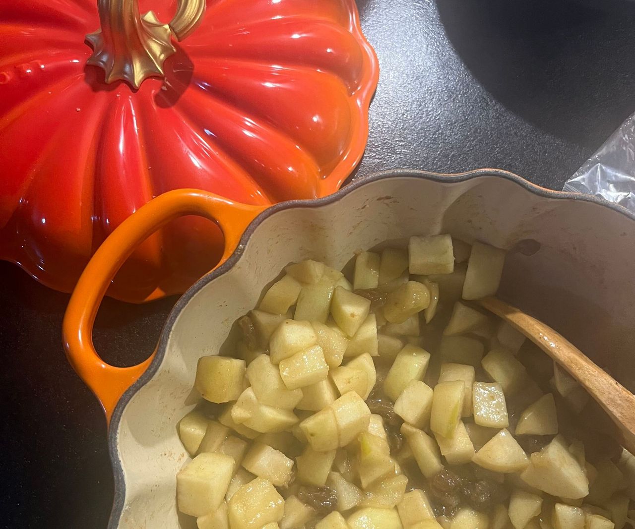 An orange Le Creuset Pumpkin cocotte on a black counter with strewed apple inside, beside its pumpkin shaped lid.