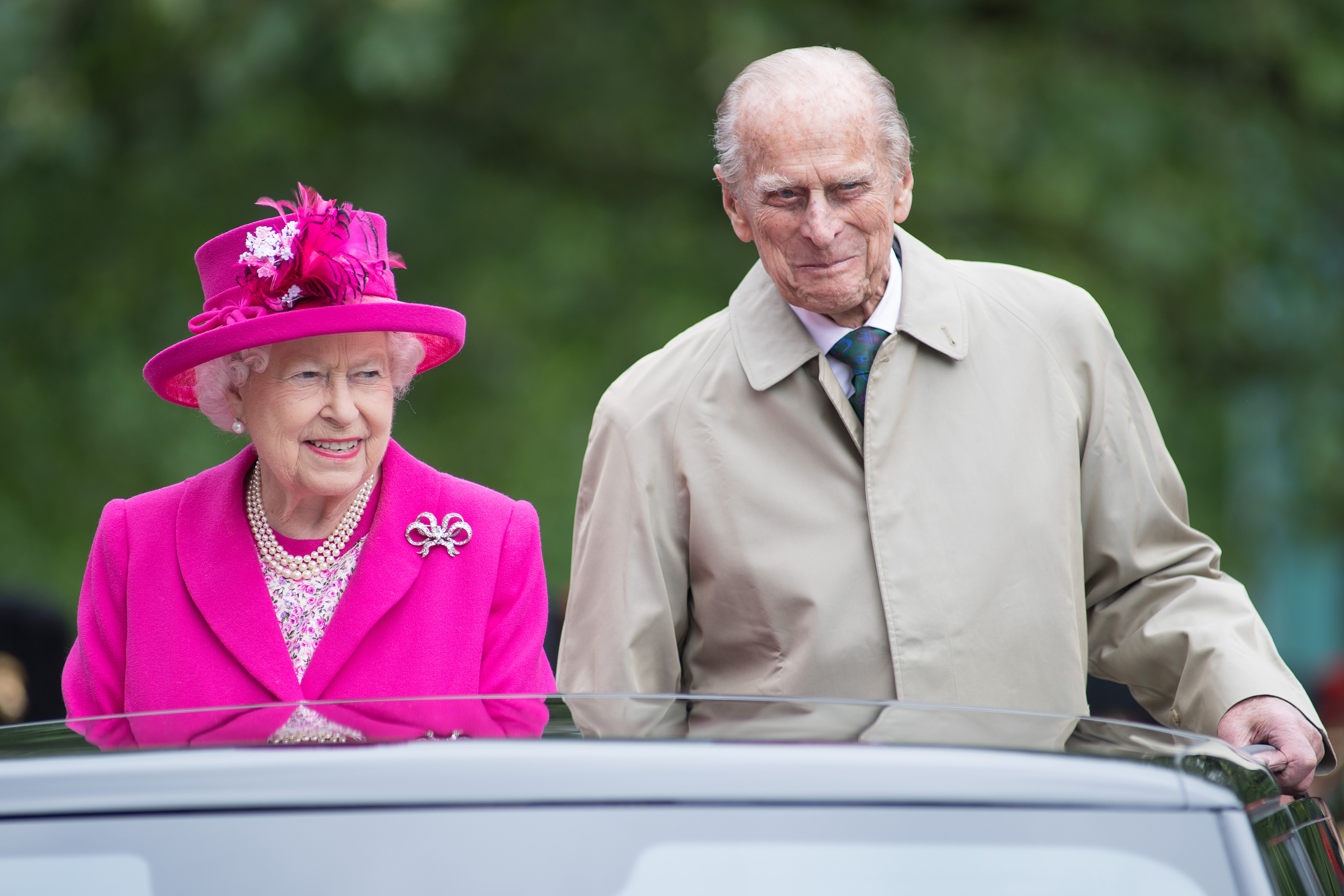Queen Elizabeth wearing a pink coat standing out the sunroof of a car with Prince Philip
