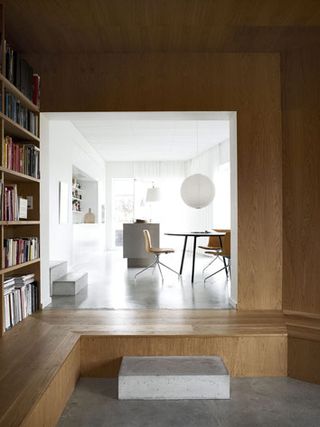From the oak-clad living room, through an opening, we see a white and very bright concrete kitchen. We see a table with three yellow chairs.
