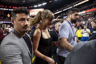 Taylor Swift being escorted by a bodyguard in a gray suit in a crowd across the field at the 2024 Super Bowl