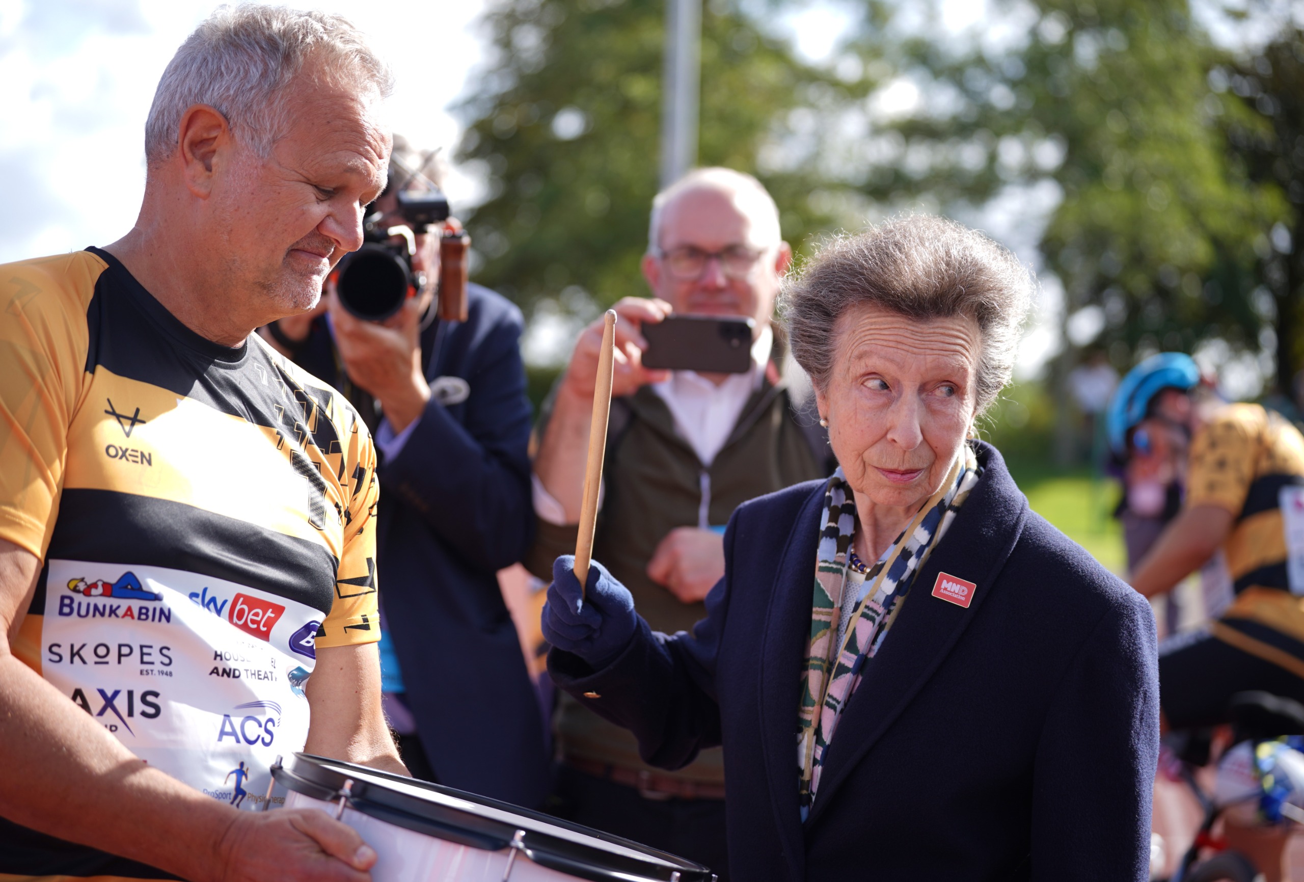 Princess Anne banging a drum next to a man in a yellow jersey