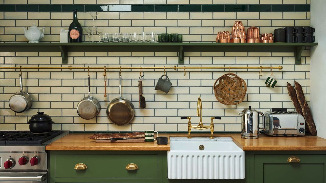 A white subway-tiled kitchen with green cabinets and open wall shelving. There are copper and stainless steel pans on display on a brass hanging rail above the white undermounted butler sink