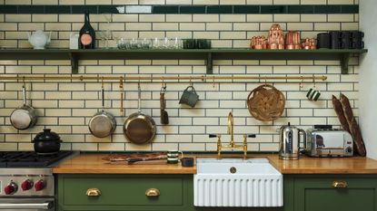 A white subway-tiled kitchen with green cabinets and open wall shelving. There are copper and stainless steel pans on display on a brass hanging rail above the white undermounted butler sink