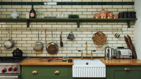 A white subway-tiled kitchen with green cabinets and open wall shelving. There are copper and stainless steel pans on display on a brass hanging rail above the white undermounted butler sink