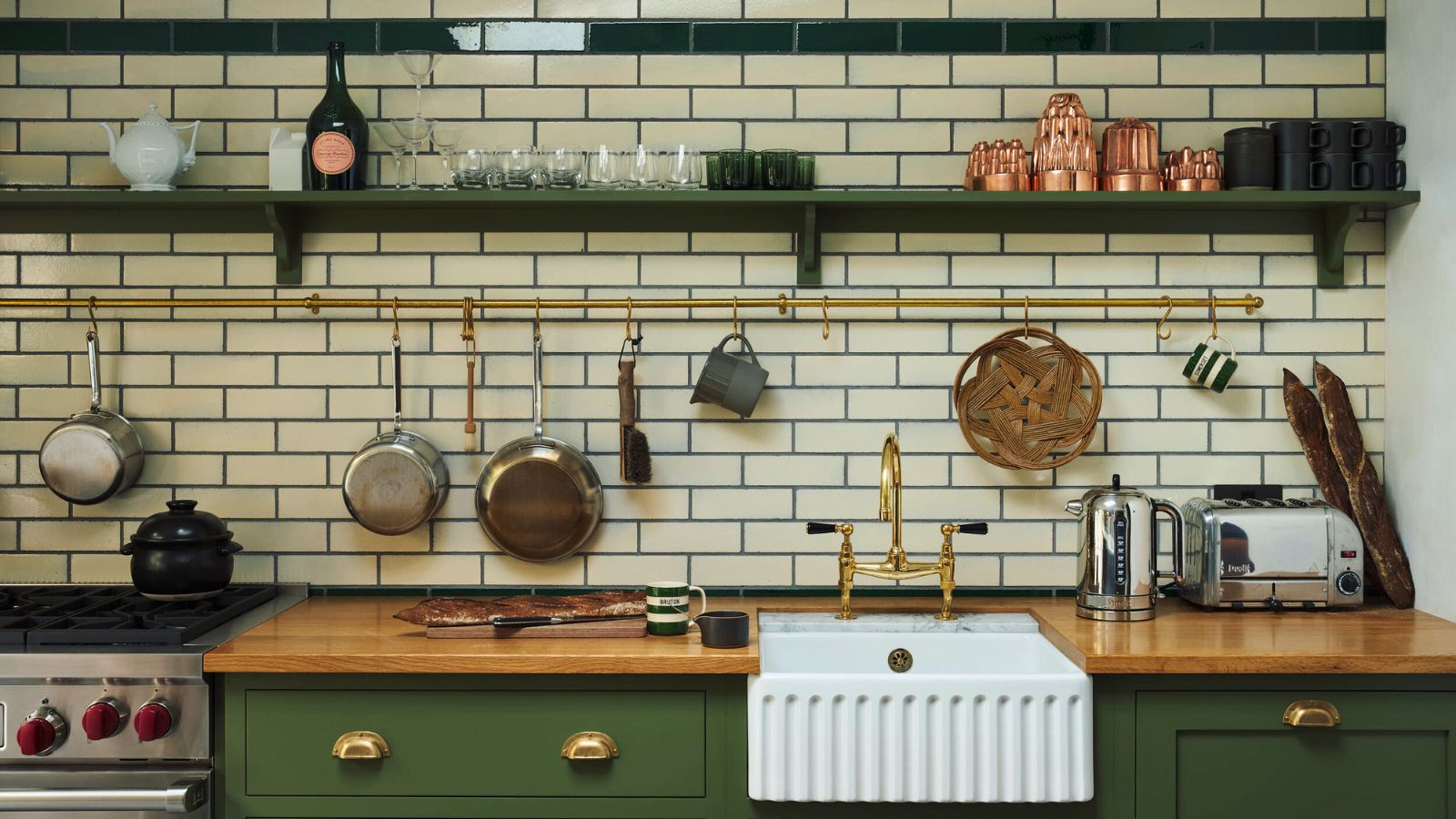 A white subway-tiled kitchen with green cabinets and open wall shelving. There are copper and stainless steel pans on display on a brass hanging rail above the white undermounted butler sink
