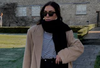 Influencer wears camel coat, striped shirt, black jeans, black scarf, and sunglasses.