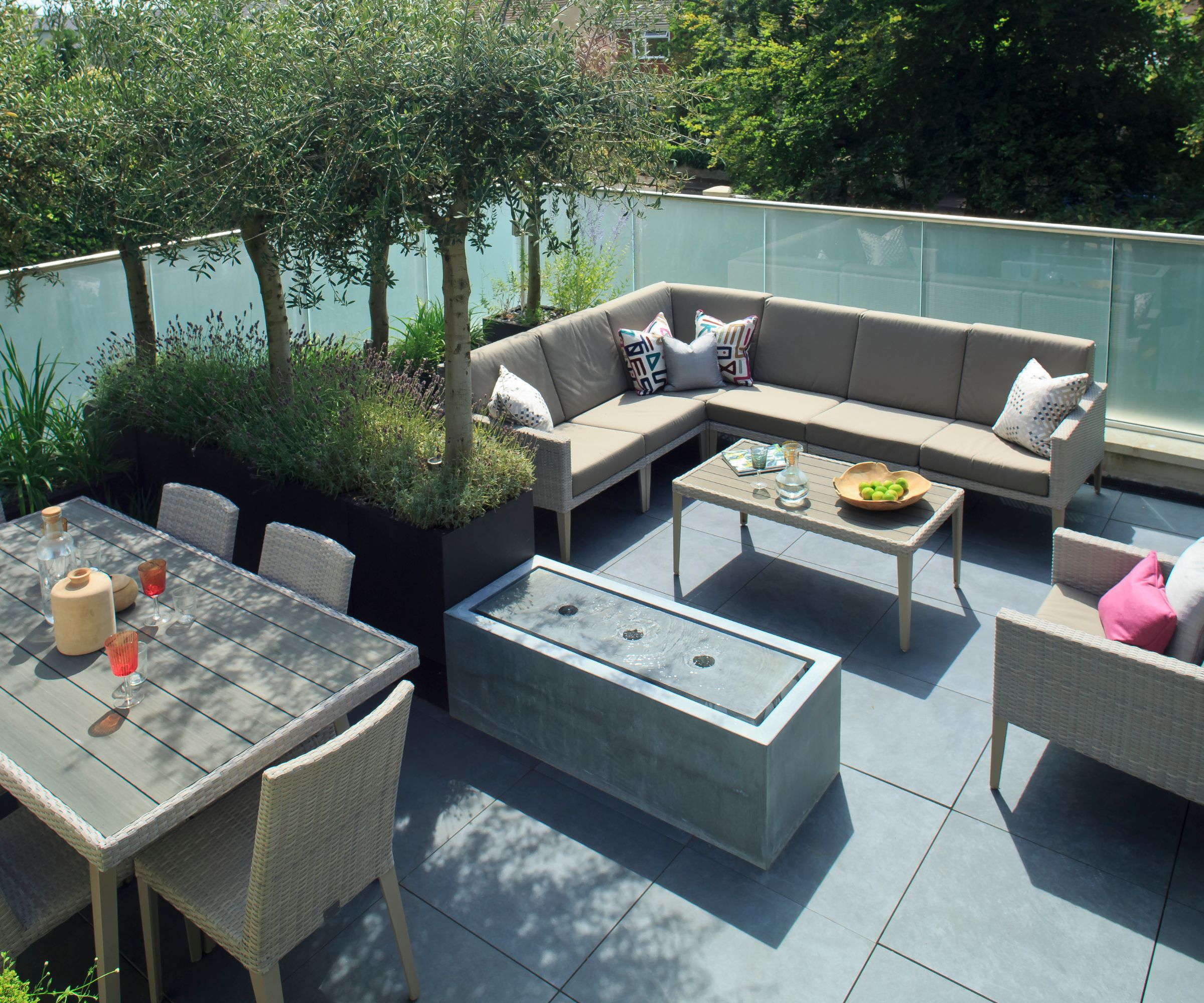 Rooftop terrace with corner sofa, dining table, water feature, and potted olive trees behind glass balustrade.