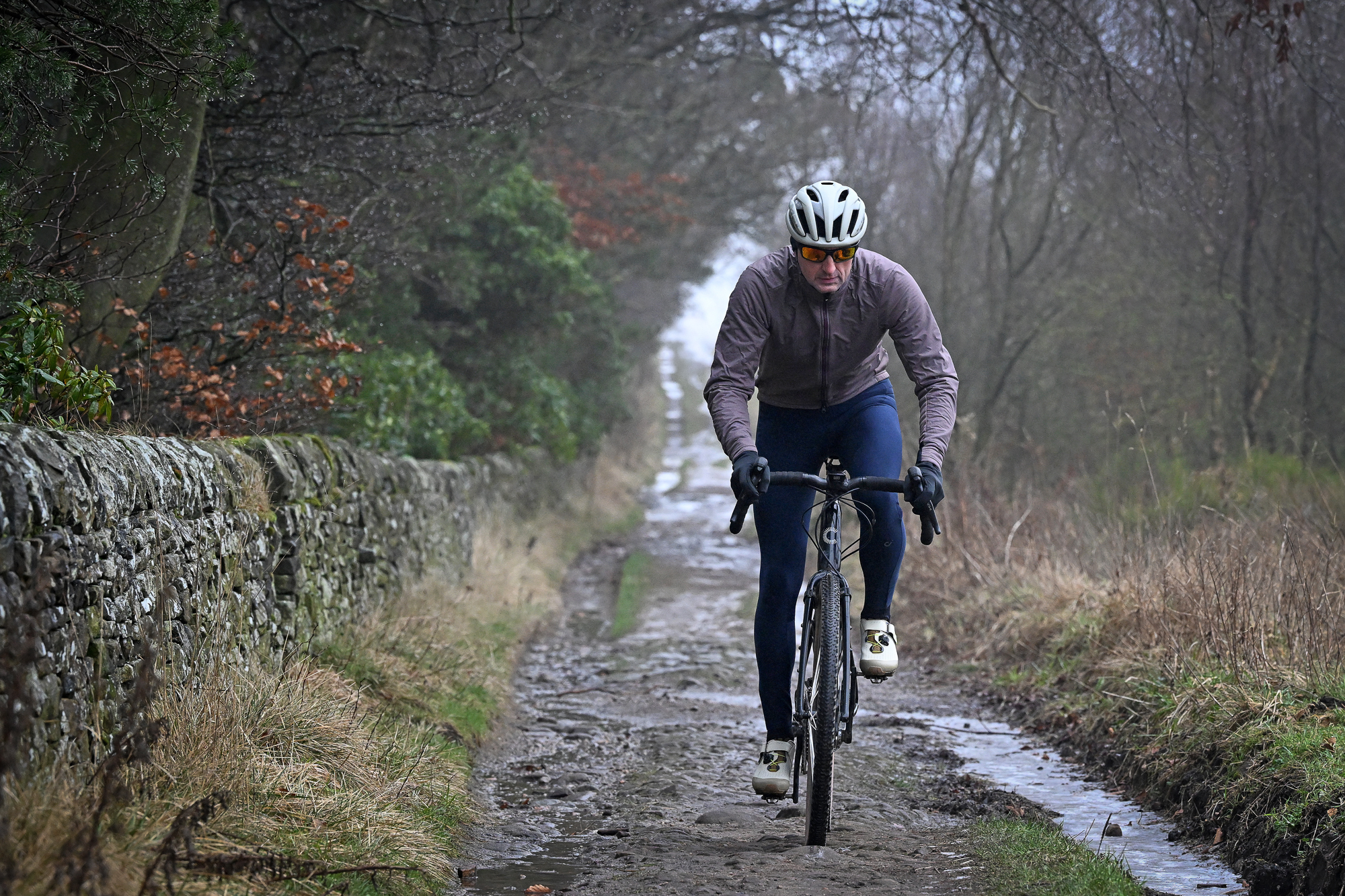 Man wearing a pale purple jacket riding a gravel bike towards the camera on a wet trail