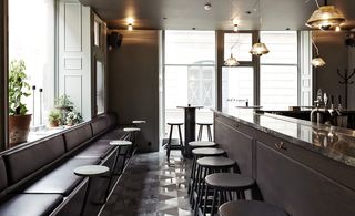 The Flying Elk, Stockholm, Sweden. A bar with a dark wood marble topped counter with round high chairs and a leather sitting bench along the wall with round tables in front of it.