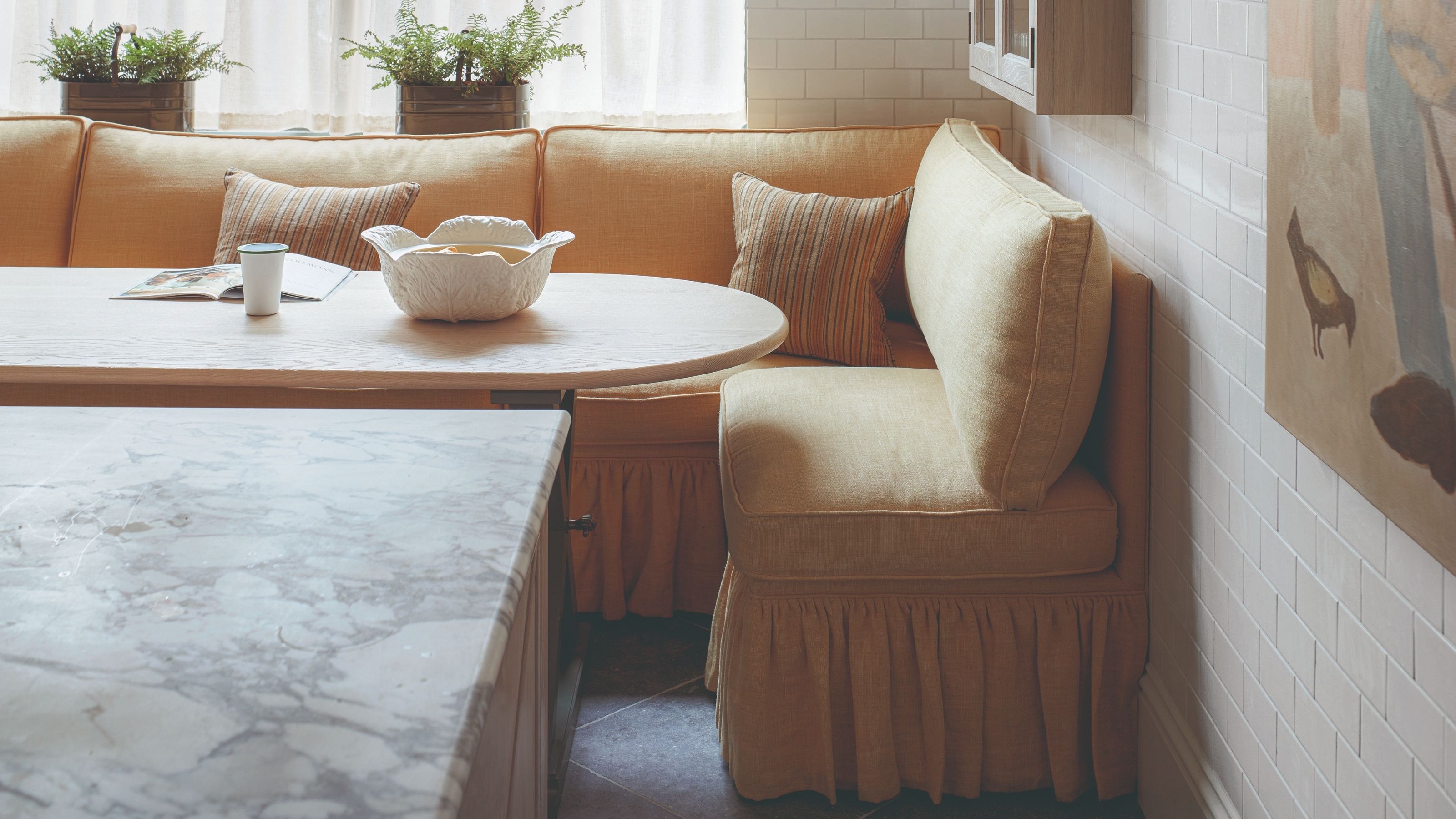 A yellow ruffled banquette seat in a breakfast nook with a curved table. The walls are white tile, and there is art and plants styled around it.