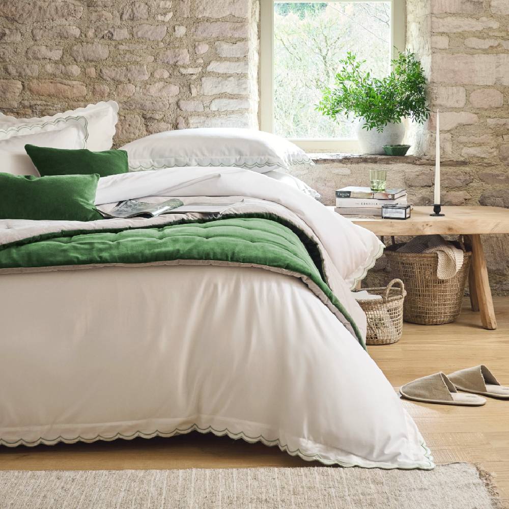 A rustic bedroom with a stone wall, with a luxurious bedspread from Christy, with slippers and a wooden bedside table.