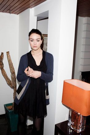 Female model, wearing a dark blue detailed cardigan, black dress and tights, orange lamp, white walls, dark wood ceiling, black gloss side table, potted plant