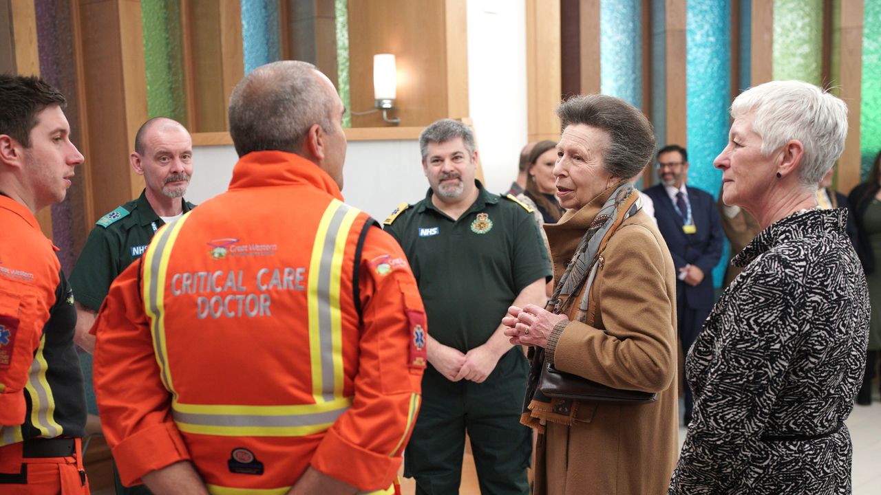 Princess Anne wearing a brown coat standing and talking with doctors at a hospital including a man with an orange coat reading &quot;critical care doctor&quot;