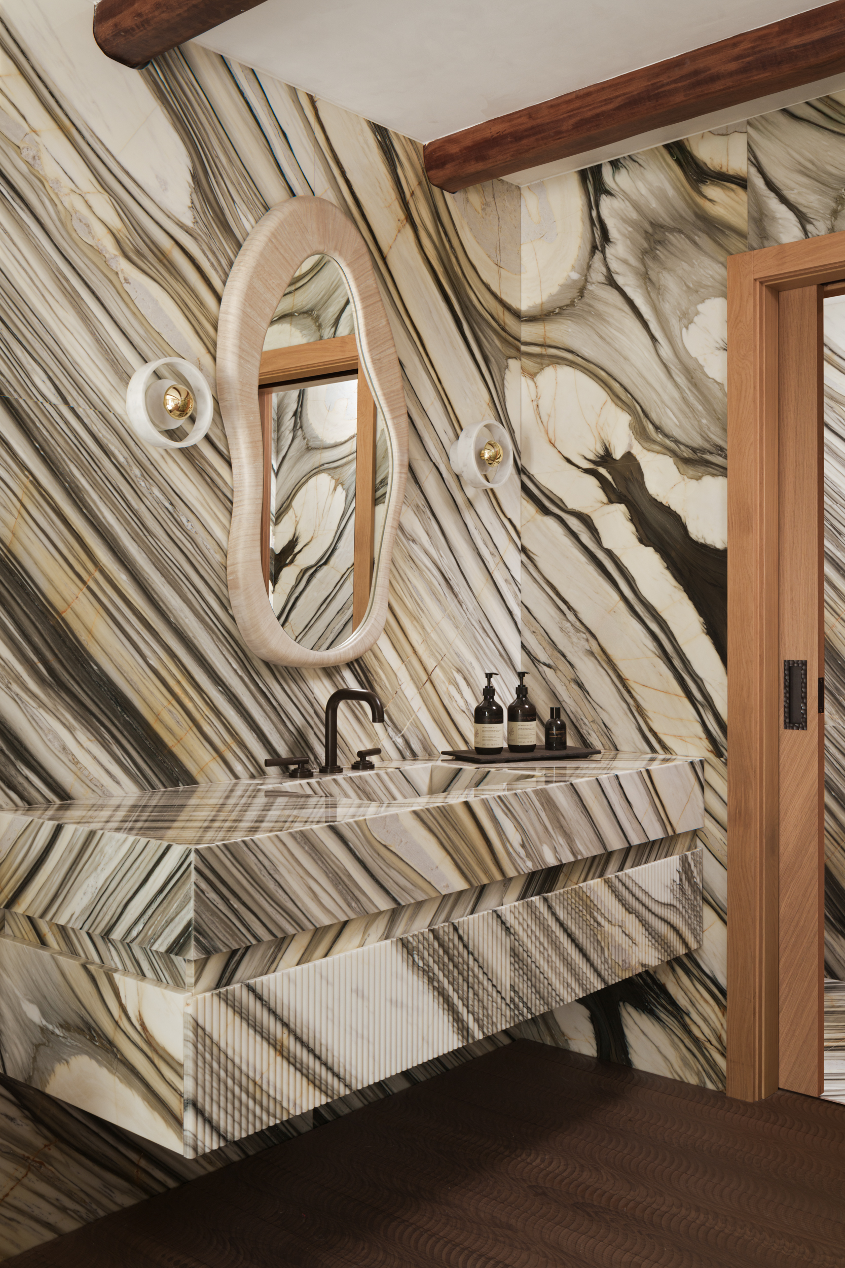 A bathroom with marble walls and vanity. and an organic shaped mirror