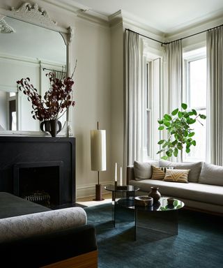 A white living room with high ceilings, a dark blue area rug, a large mirror above a fireplace, and a glass coffee table