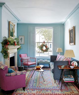 Living room aqua walls, high ceiling, cornice, fireplace with mantelpiece, sash window with wooden shutters, floor rug and colourful cushions on soft furnishings.