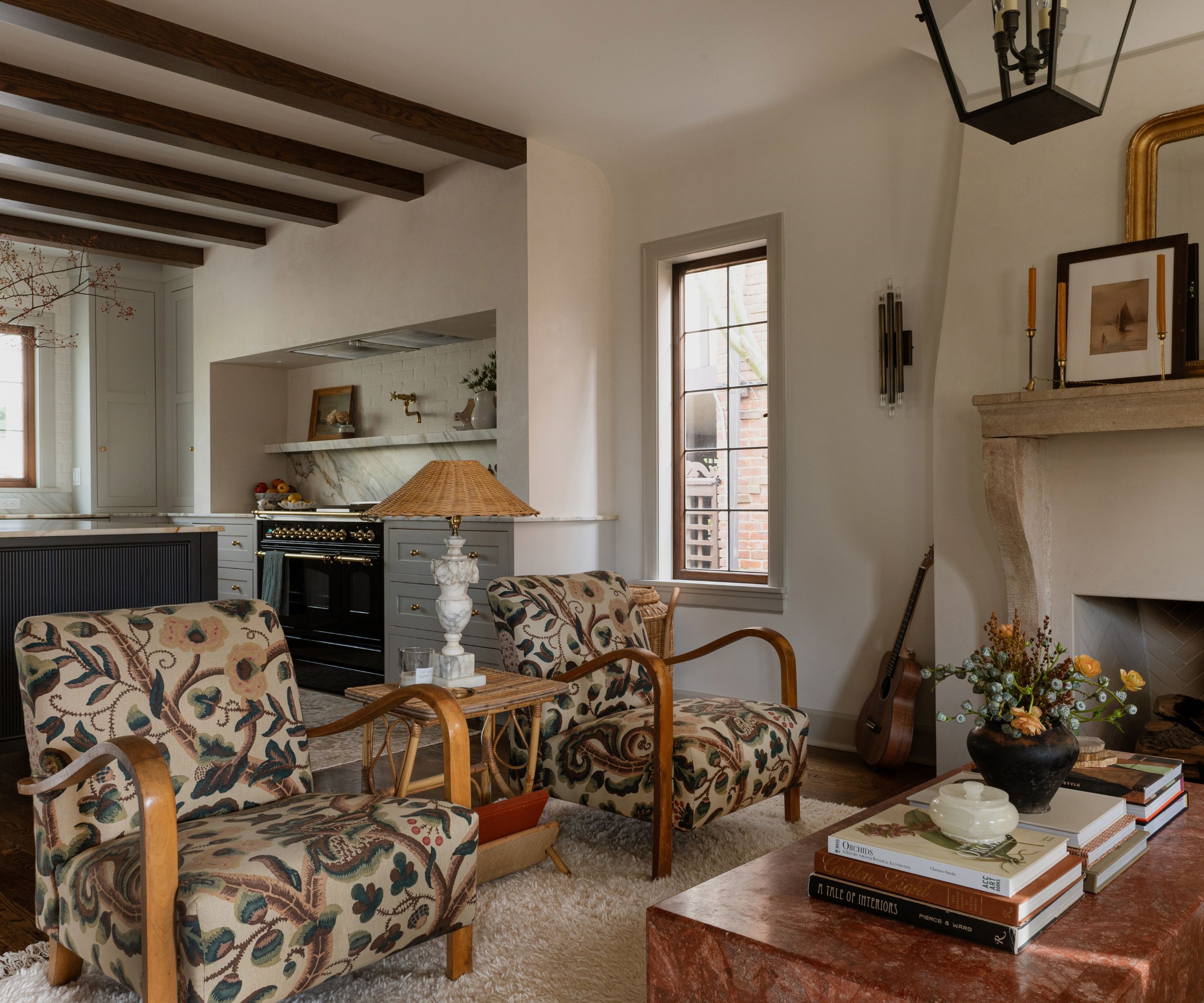 Living area in an open plan kitchen with stone mantle, vintage chairs upholstered in a vintage floral print, stone coffee table decorated with coffee table books