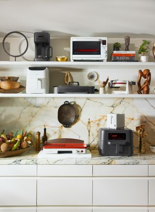 CruxGG kitchenware collection including coffee maker, air fryer and grill displayed on a light marble worksurface and white shelves with ornaments, small plants and other kitchenware throughout