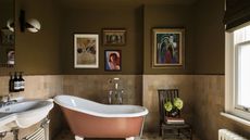 Bathroom with dark olive painted walls, half tiled in cream tiles, a terracotta coloured freestanding tub, artwork on the wall, and a white sink.