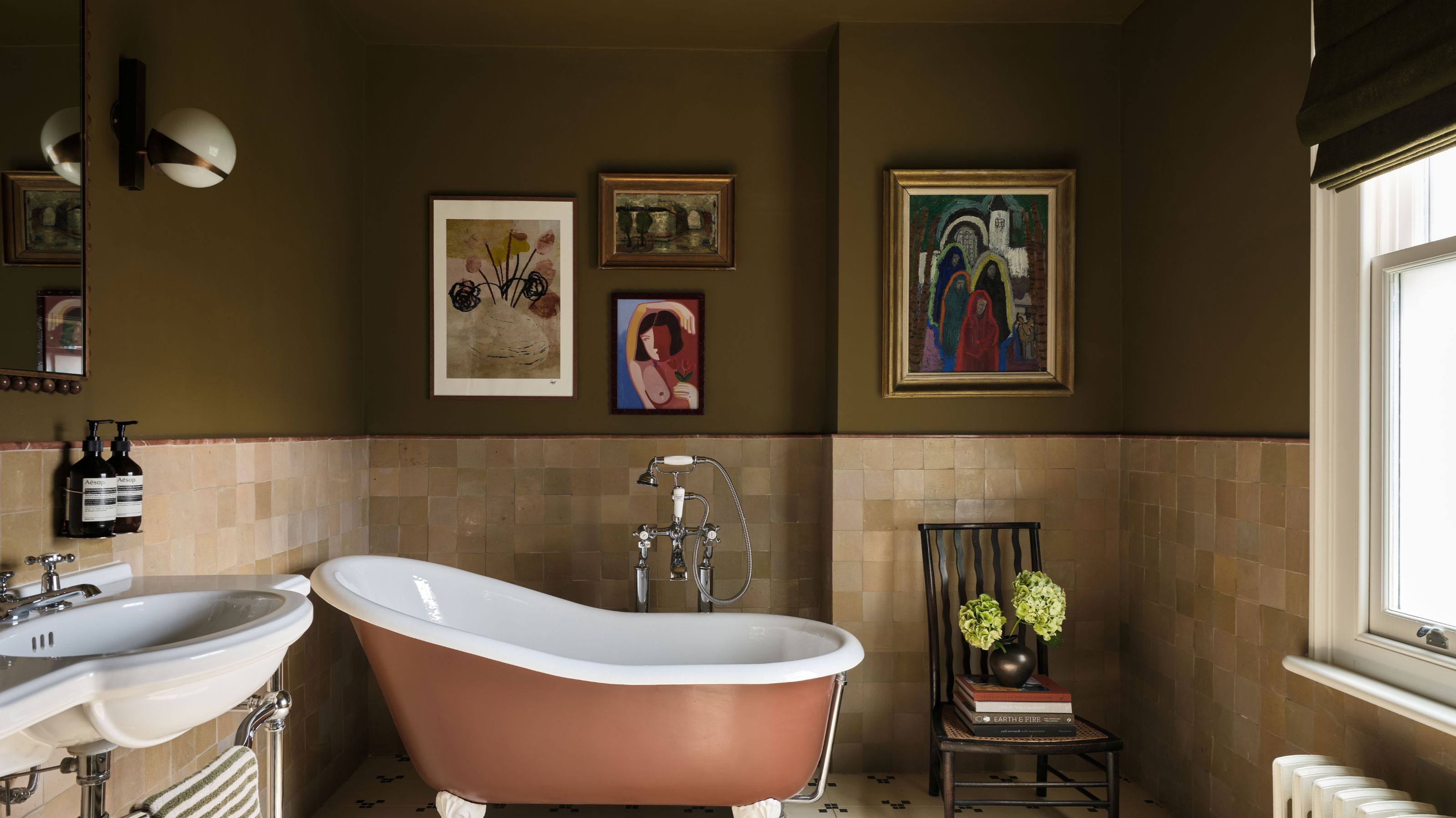 Bathroom with dark olive painted walls, half tiled in cream tiles, a terracotta coloured freestanding tub, artwork on the wall, and a white sink.
