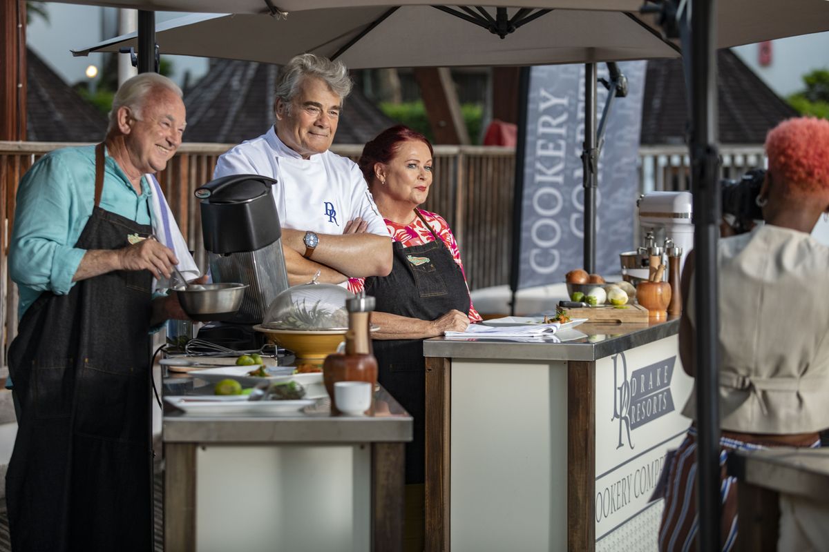 A still from Death In Paradise season 13 episode 3 showing Michael Fenton Stevens as Tristan Clayborn, Gordon Kennedy as Stanley Drake and Kate Robbins as Lucky Clayborn, all standing behind their counters at a chef competition posing for a photo being taken by Andrina Harper (Genesis Lynea)