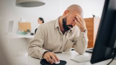 Visibly stressed male office worker with fingers on temples while working on a desktop computer.
