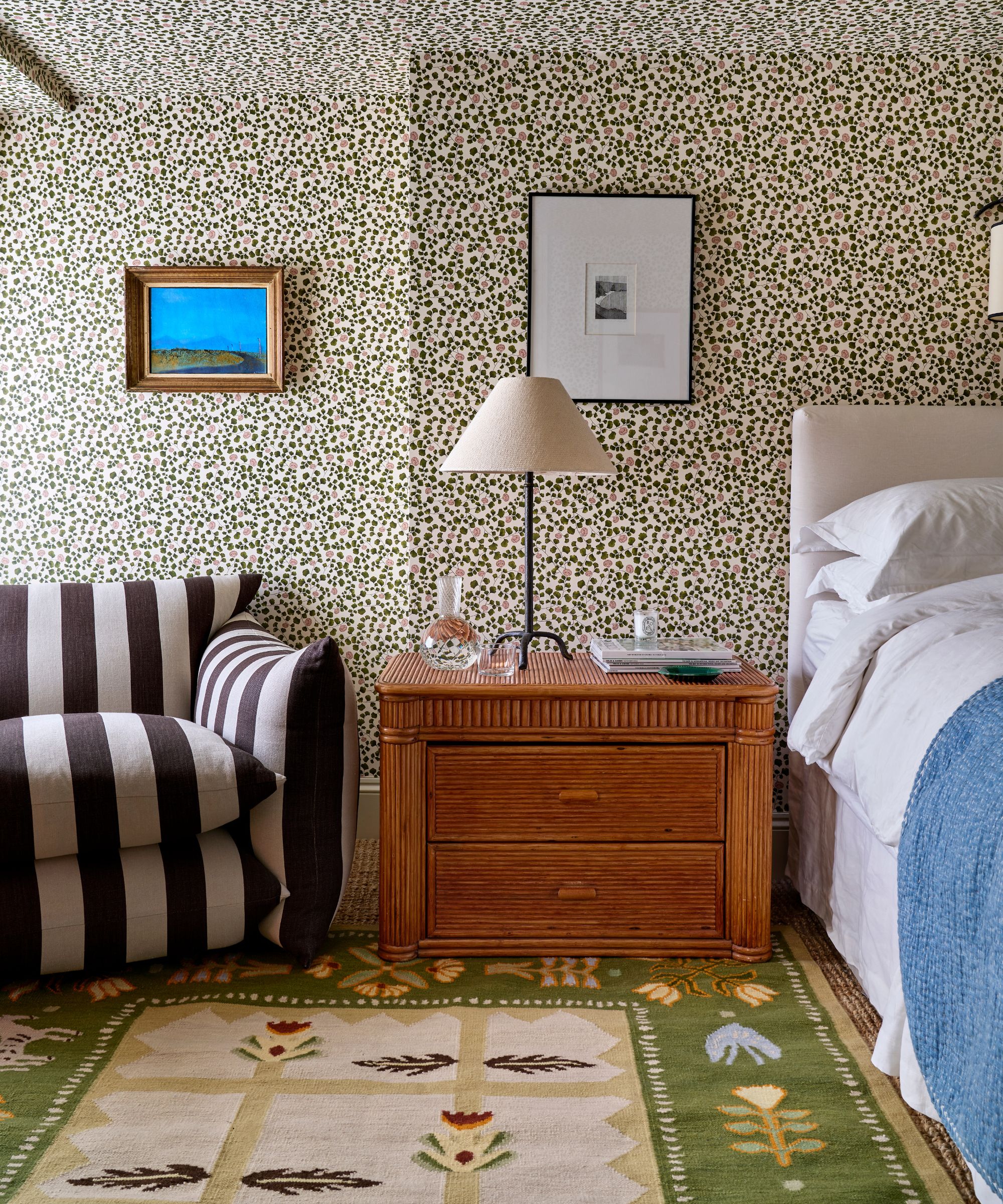 guest attic bedroom with floral wallpapered walls and ceiling with a flatweave swedish style green floral rug, a wooden vintage nightstand, with a black and white striped arm chair