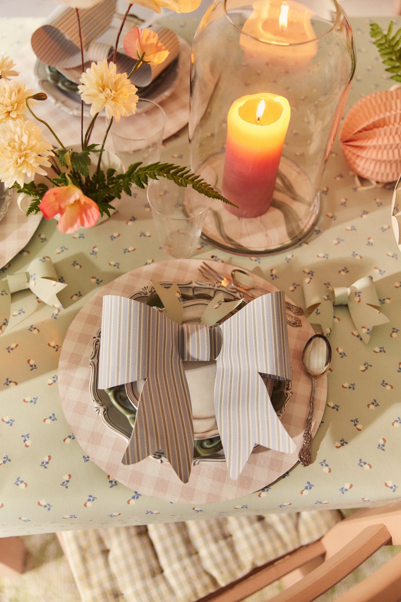 Image of a pastel table set for a spring dinner party. There is a light mint green wallpaper as a tablecloth and a pink gingham placemat. There is a blue and white striped bow made out of wallpaper on top of the plate.