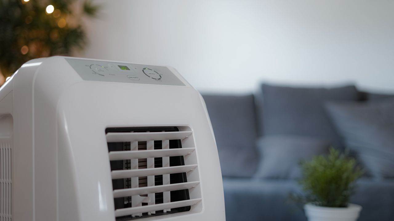 Portable air conditioner unit in living room, in front of a grey sofa, plant and white wall