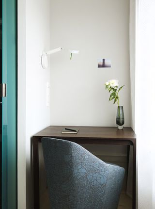 Hotel Skeppsholmen, Stockholm. A small dark wood desk with a book and pen on it, a vase with a white flower in it, a wall lamp above it and a grey fabric chair in front of it.