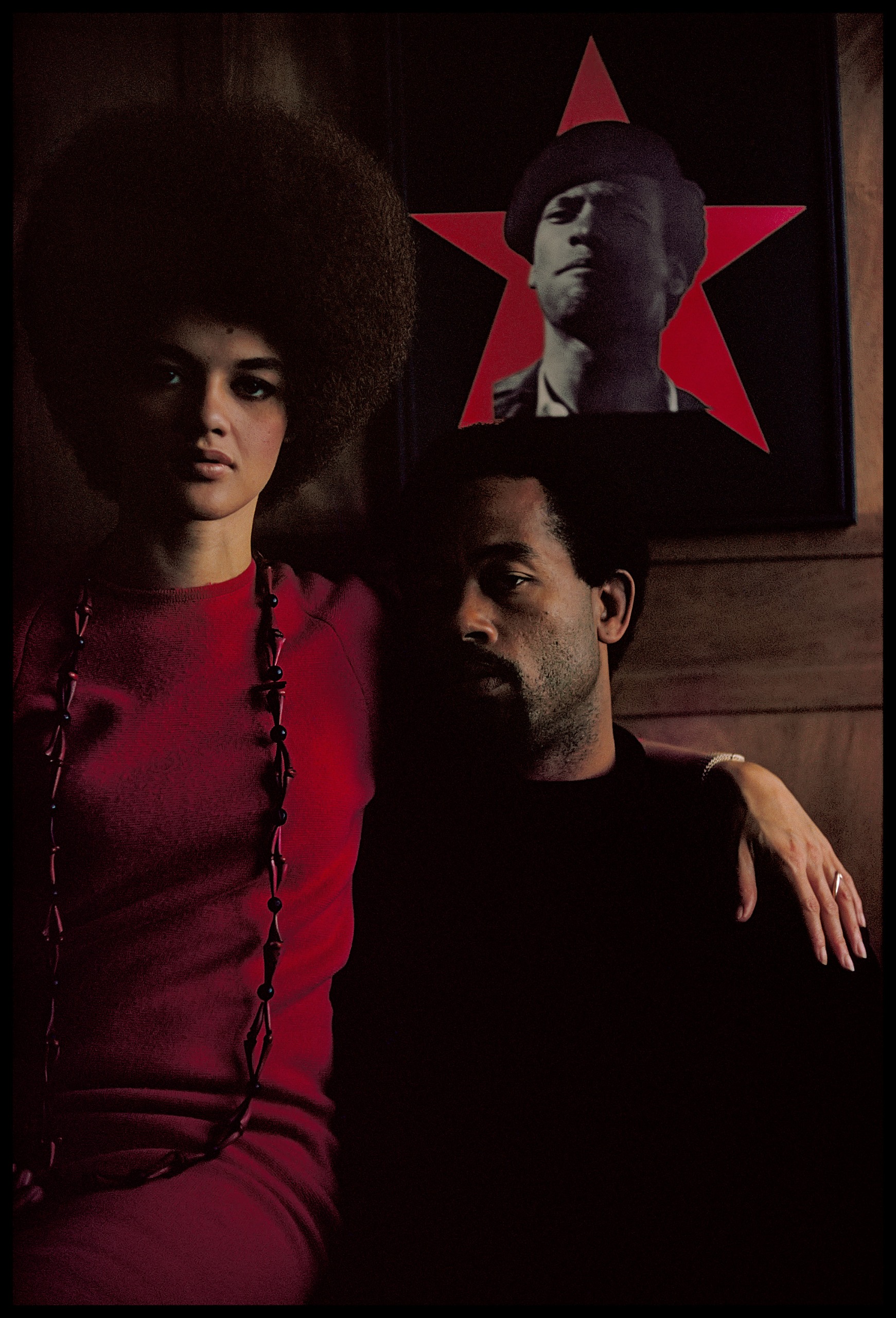 Kathleen Cleaver in a red top and beaded necklace sits beside Eldridge Cleaver, who looks away from the camera beneath a framed poster of a man's silhouette set against a red star.